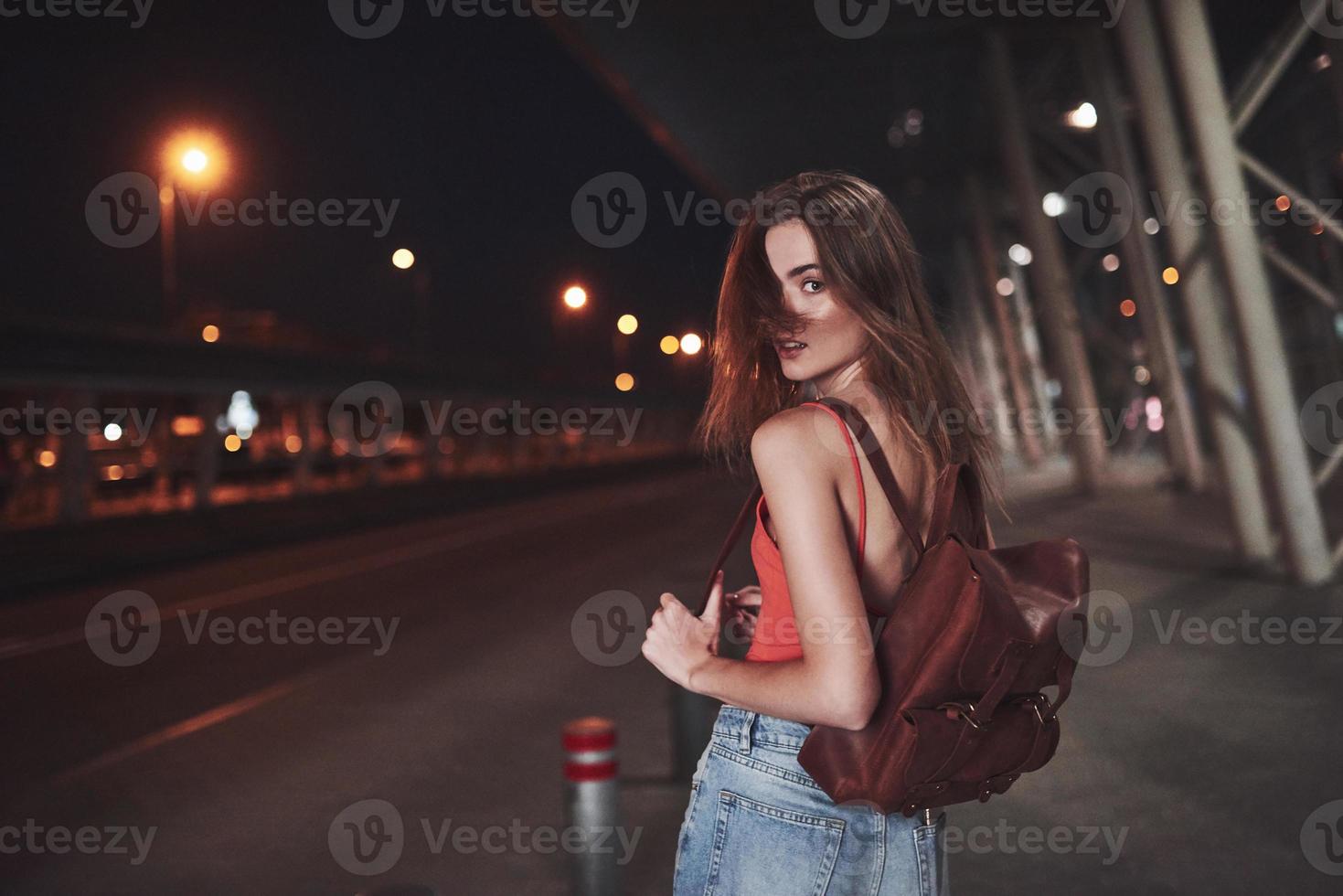 una hermosa joven con una mochila detrás de los hombros se encuentra en la calle cerca de un aeropuerto o una estación de tren en una cálida noche de verano. ella acaba de llegar y espera un taxi o sus amigos foto