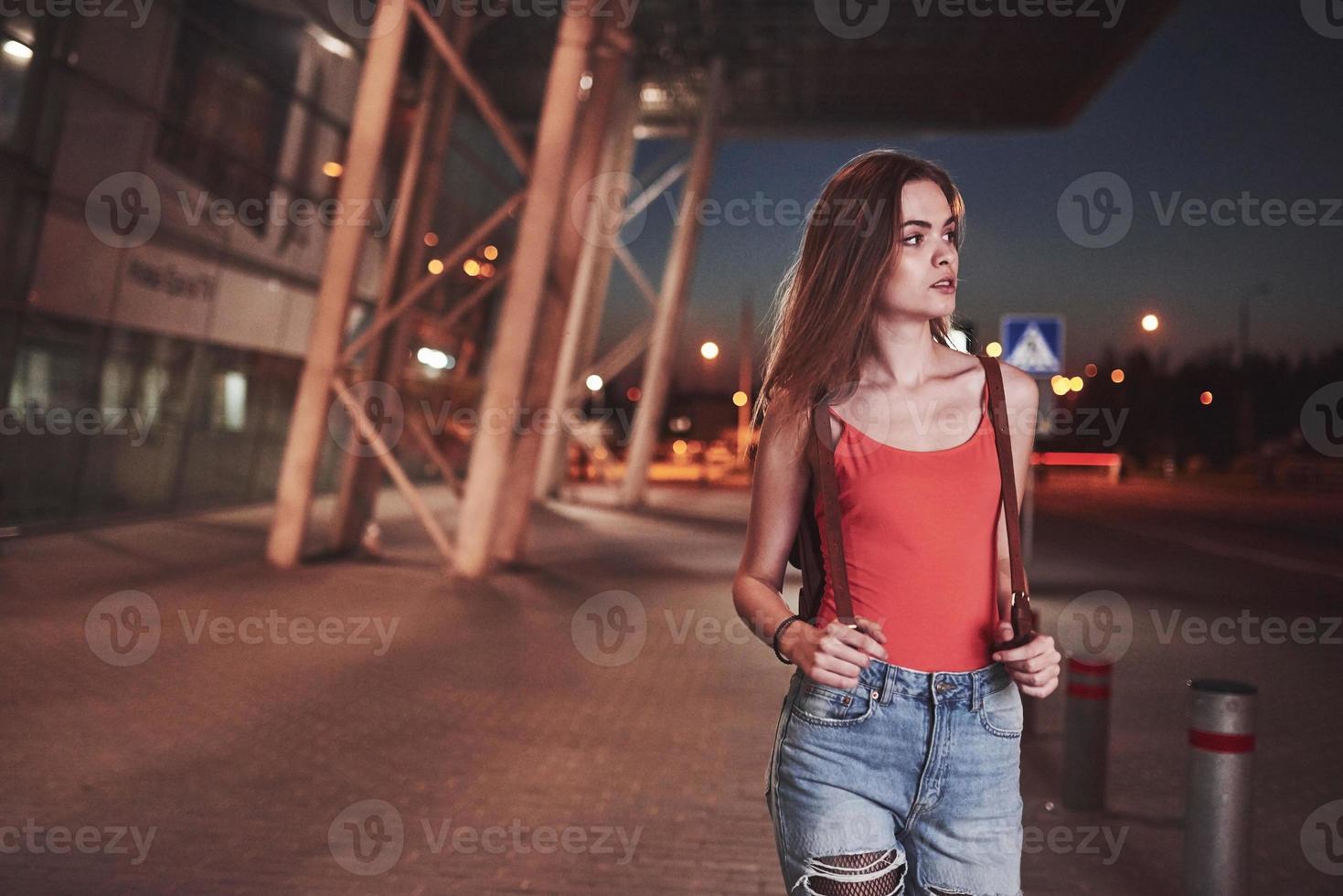 una hermosa joven con una mochila detrás de los hombros se encuentra en la calle cerca de un aeropuerto o una estación de tren en una cálida noche de verano. ella acaba de llegar y espera un taxi o sus amigos foto