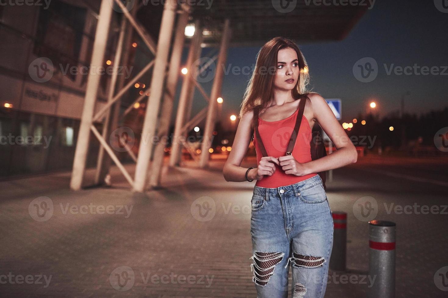 A young beautiful girl with a backpack behind her shoulders stands on the street near an airport or a railway station on a warm summer evening. She just arrived and waits for a taxi or her friends photo