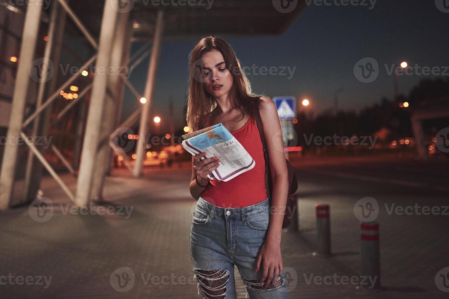 Young girl costs at night near the terminal of the airport or station and reading city map and looking for hotel. Cute tourist with backpacks determine the concept of travel photo