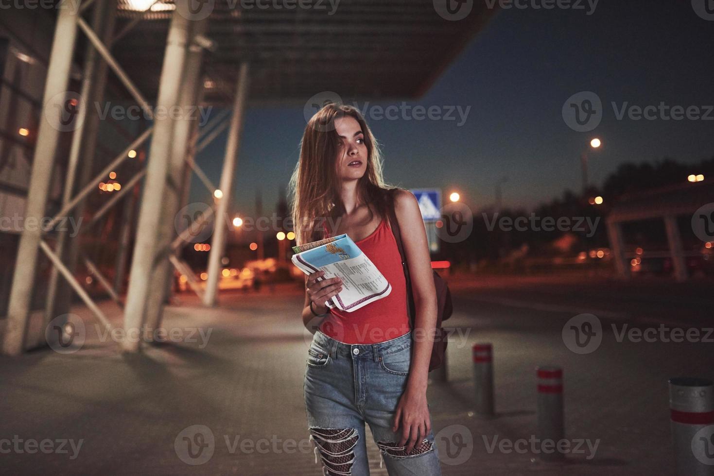 Young girl costs at night near the terminal of the airport or station and reading city map and looking for hotel. Cute tourist with backpacks determine the concept of travel photo