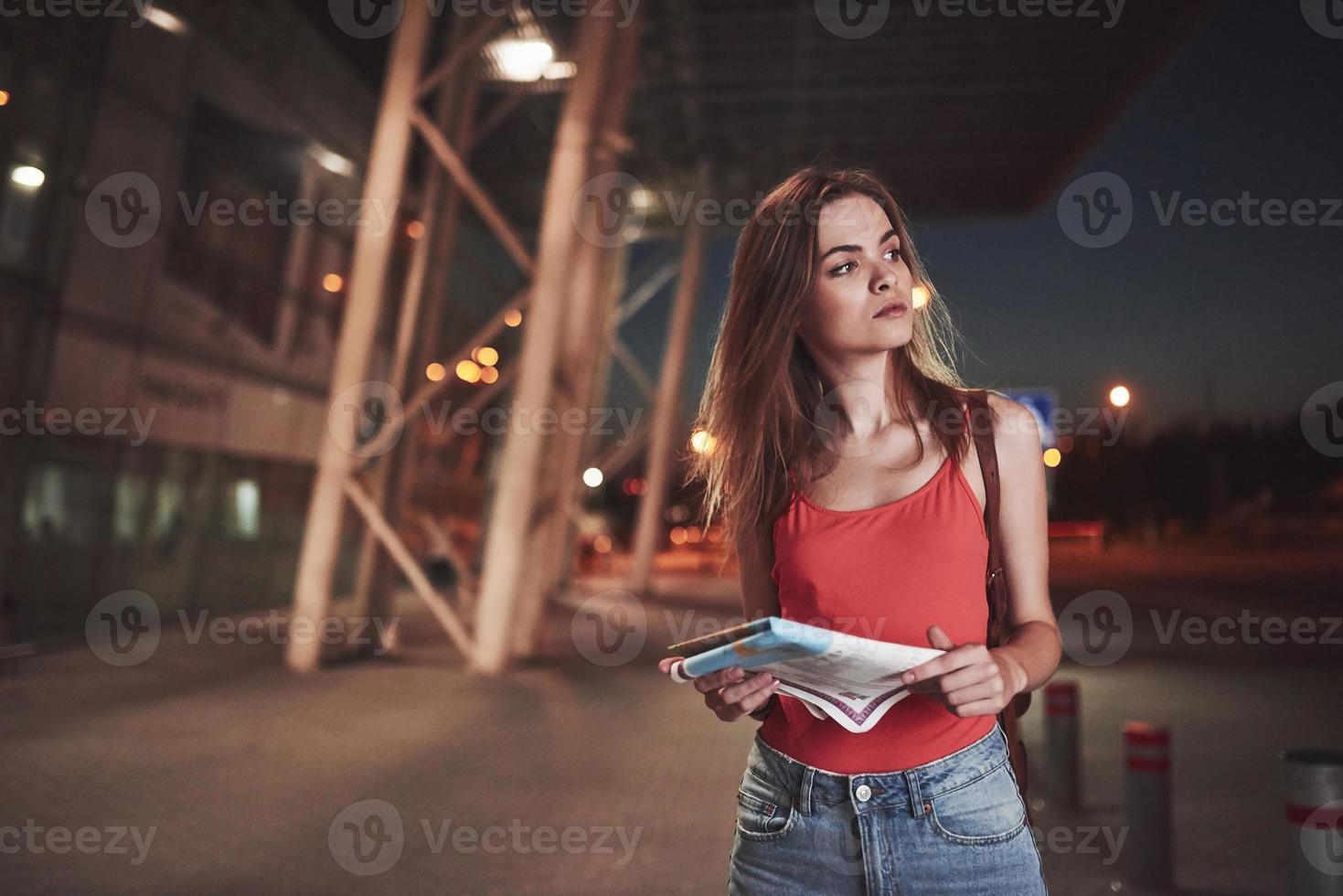 chica joven cuesta por la noche cerca de la terminal del aeropuerto o estación y leyendo mapa de la ciudad y buscando hotel. lindo turista con mochilas determinar el concepto de viaje foto