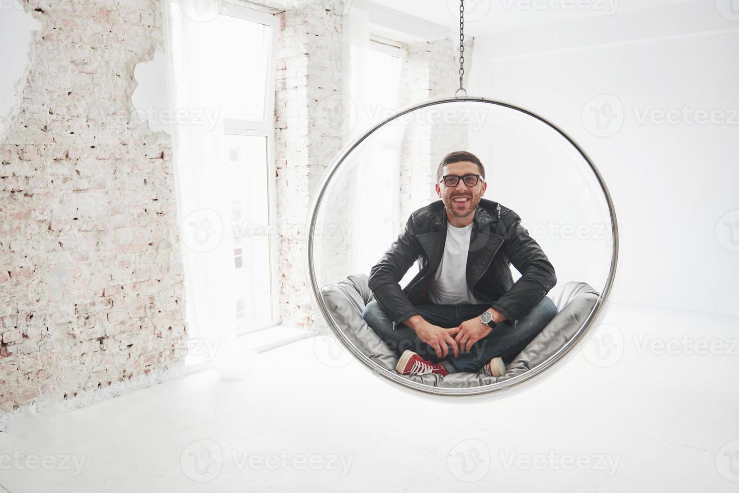 Full-length portrait of relaxed hipster man in casual sitting on hanging chair bullet and smiling on camera isolated over white background photo