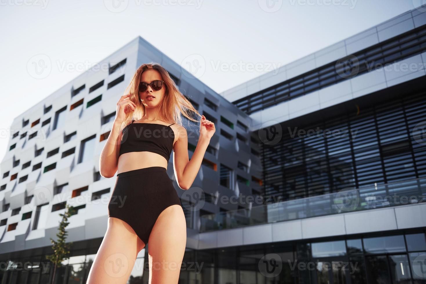 sexy mujer joven con gafas de sol y traje de baño negro está de pie en el fondo de los edificios de la ciudad durante la puesta de sol foto