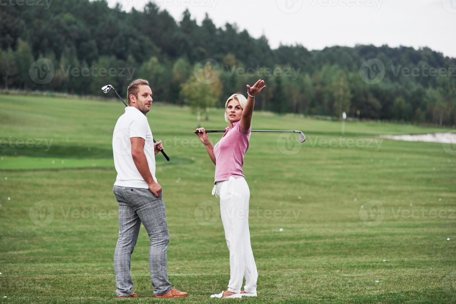 mujer mostrando algo detrás. Par de jugadores de golf con palos en sus manos de pie sobre el césped foto