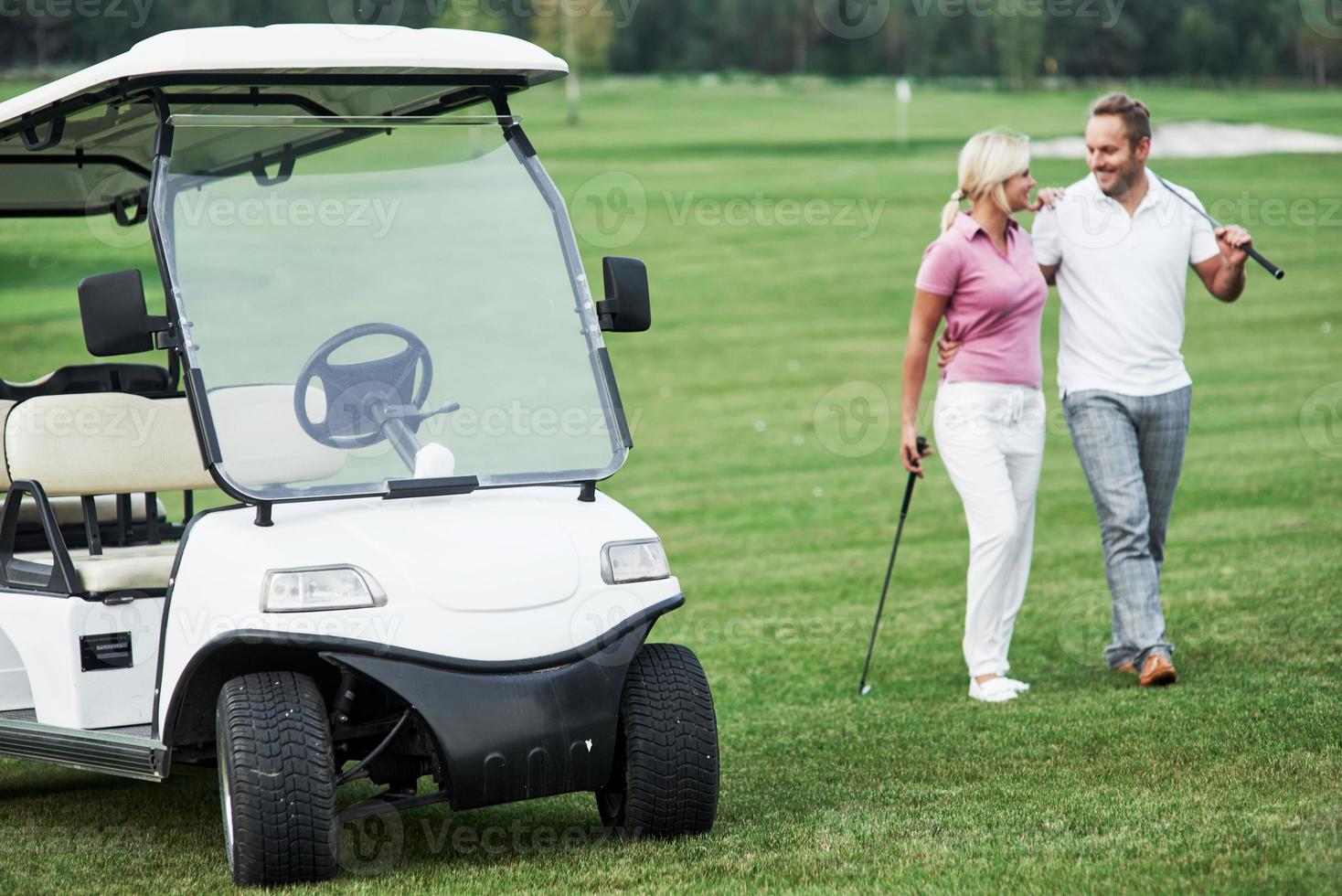 El carrito de golf con una pareja encantadora caminando cerca del vehículo y sonriendo. foto