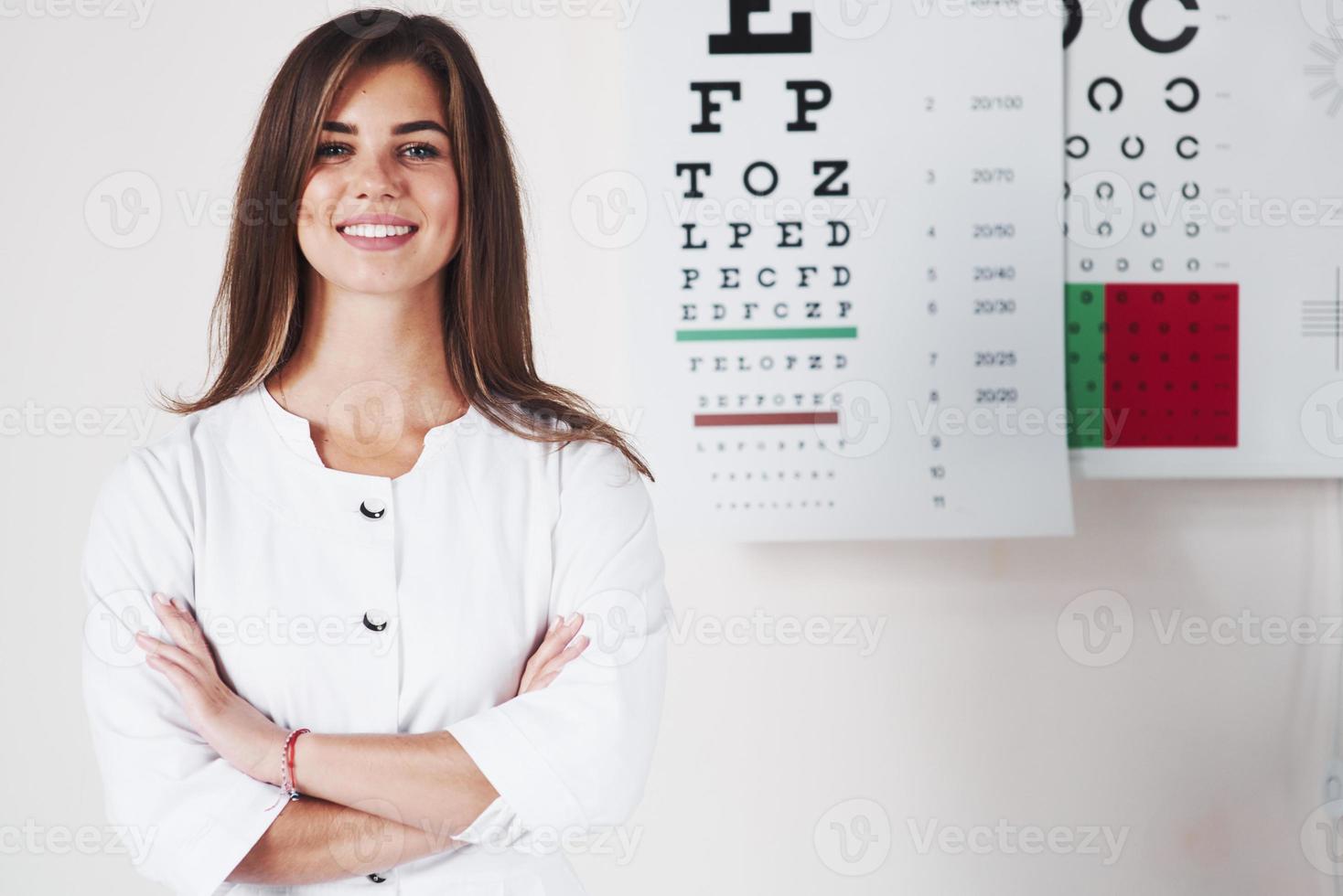 Arms crossed. Photo of gorgeous female ophthalmologist standing in the background of board with letters