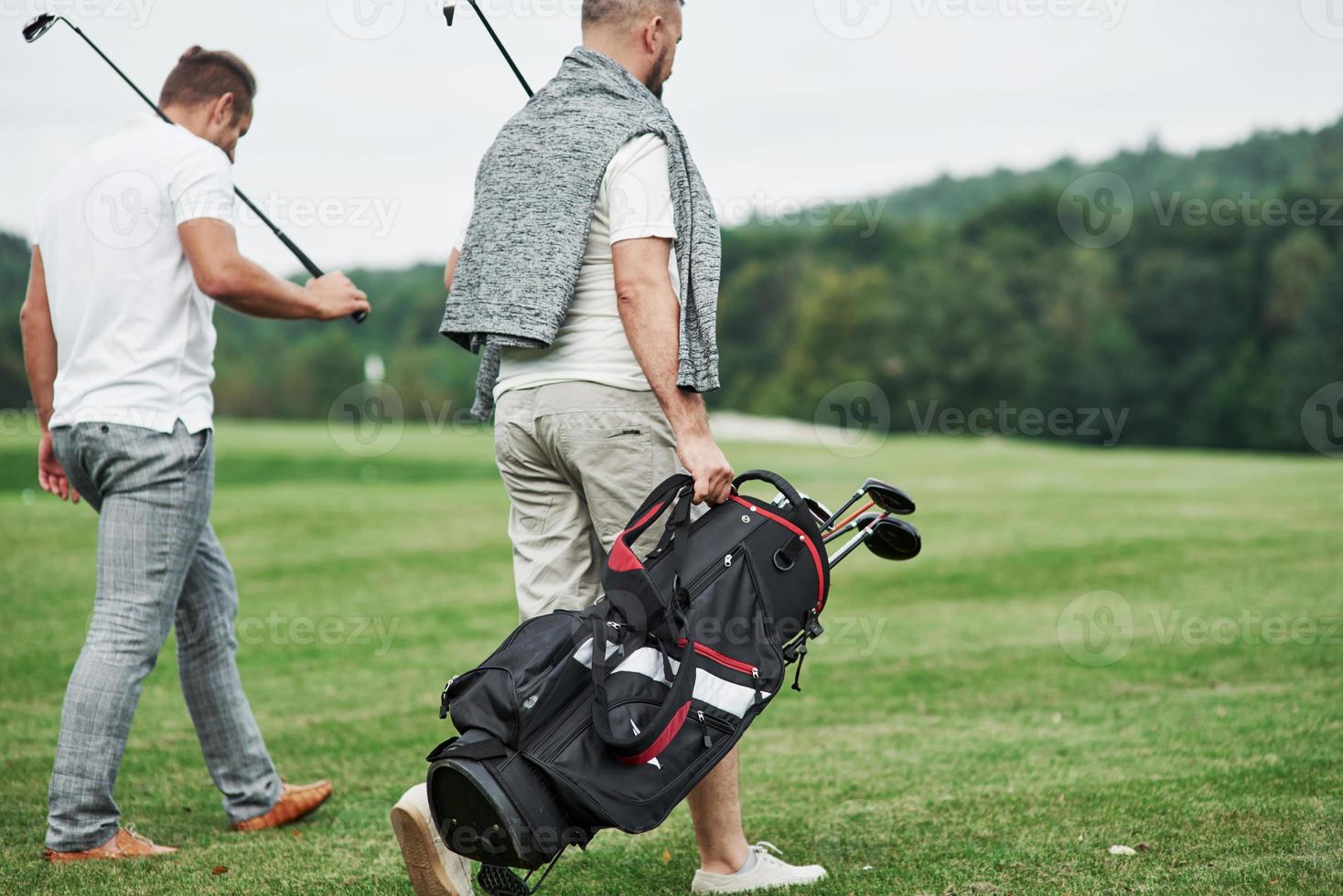 Back view. Two friends walking through the lawn with golf equipment and talking photo