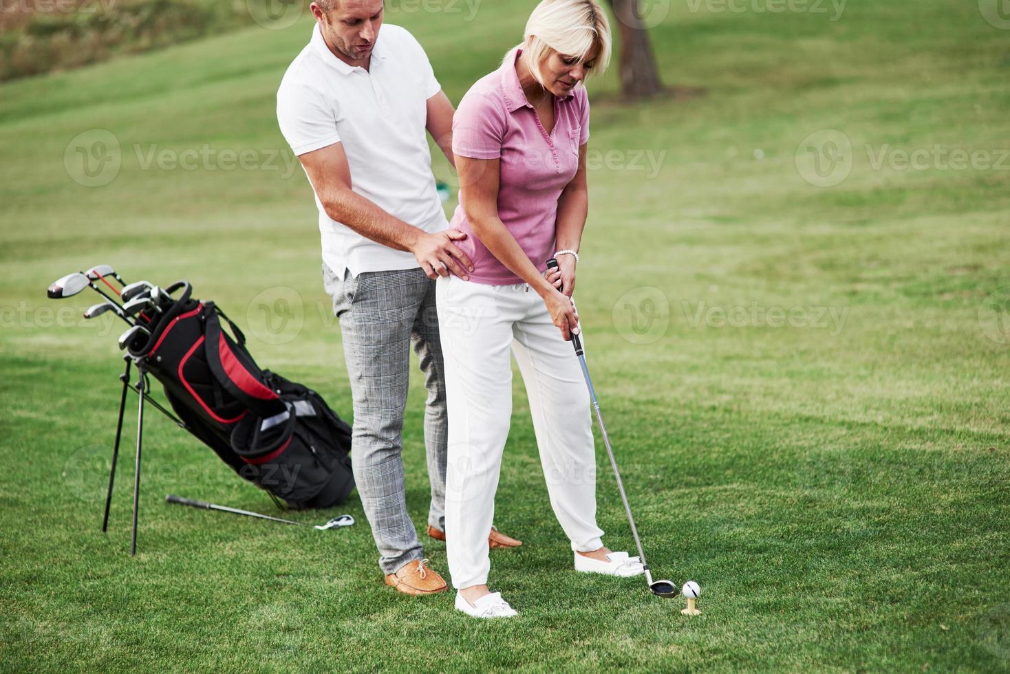 Now try on your own. Man teaching woman to play golf at the sport field with equipment behind photo