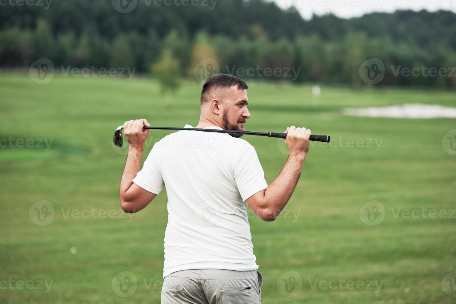 sosteniendo el palo detrás. Retrato de jugador de golf que camina en el césped y equipo en mano. bosques en el fondo foto