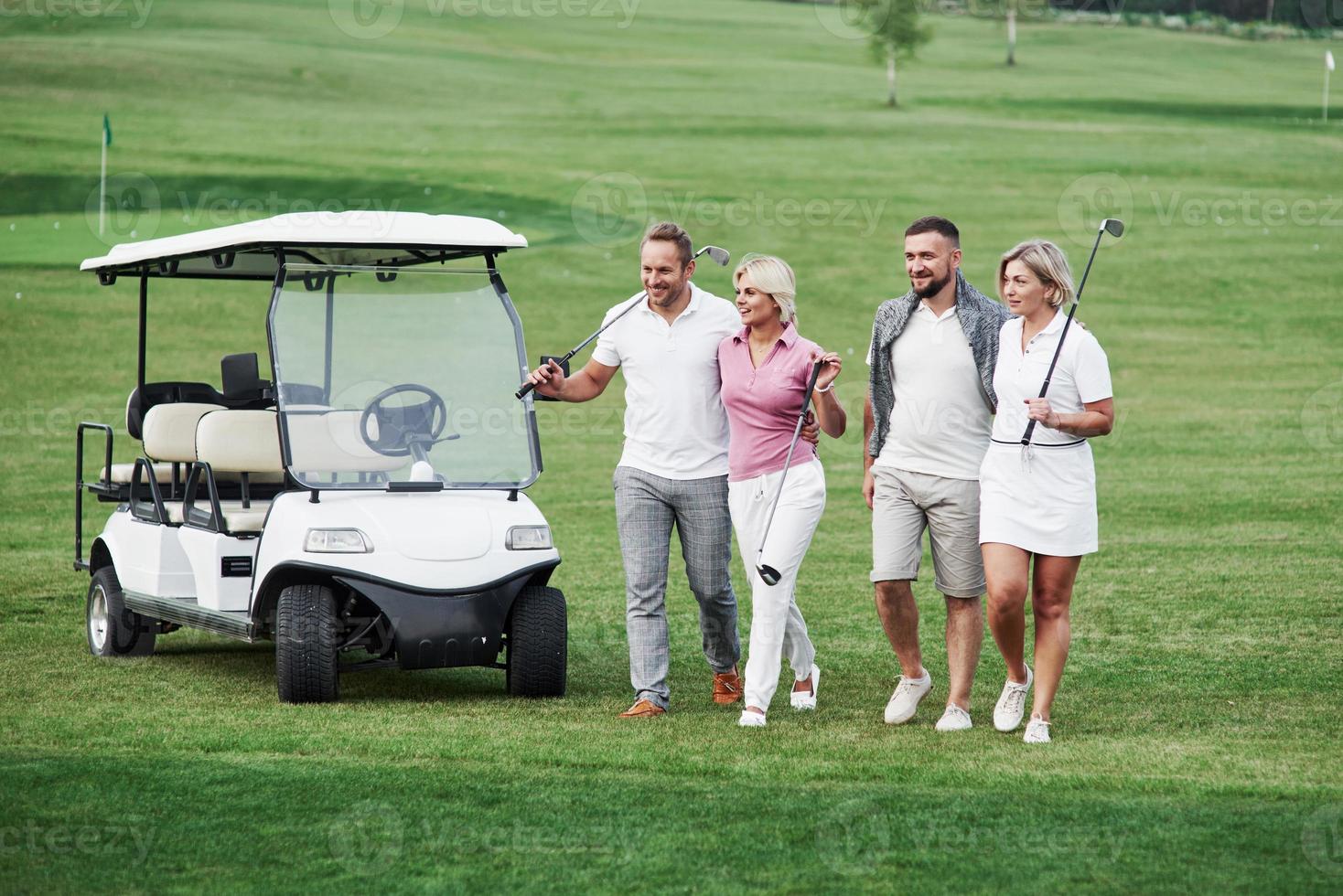 El grupo de amigos de la familia caminando por el campo después del juego de golf con el vehículo detrás. foto