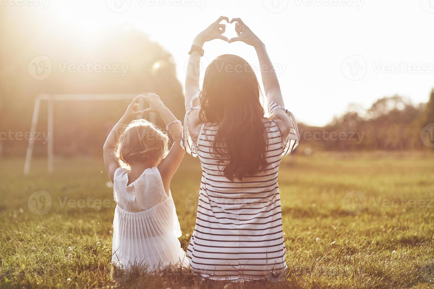 Young mother and her daughter sitting on the grass and making heart symbol to the sun photo