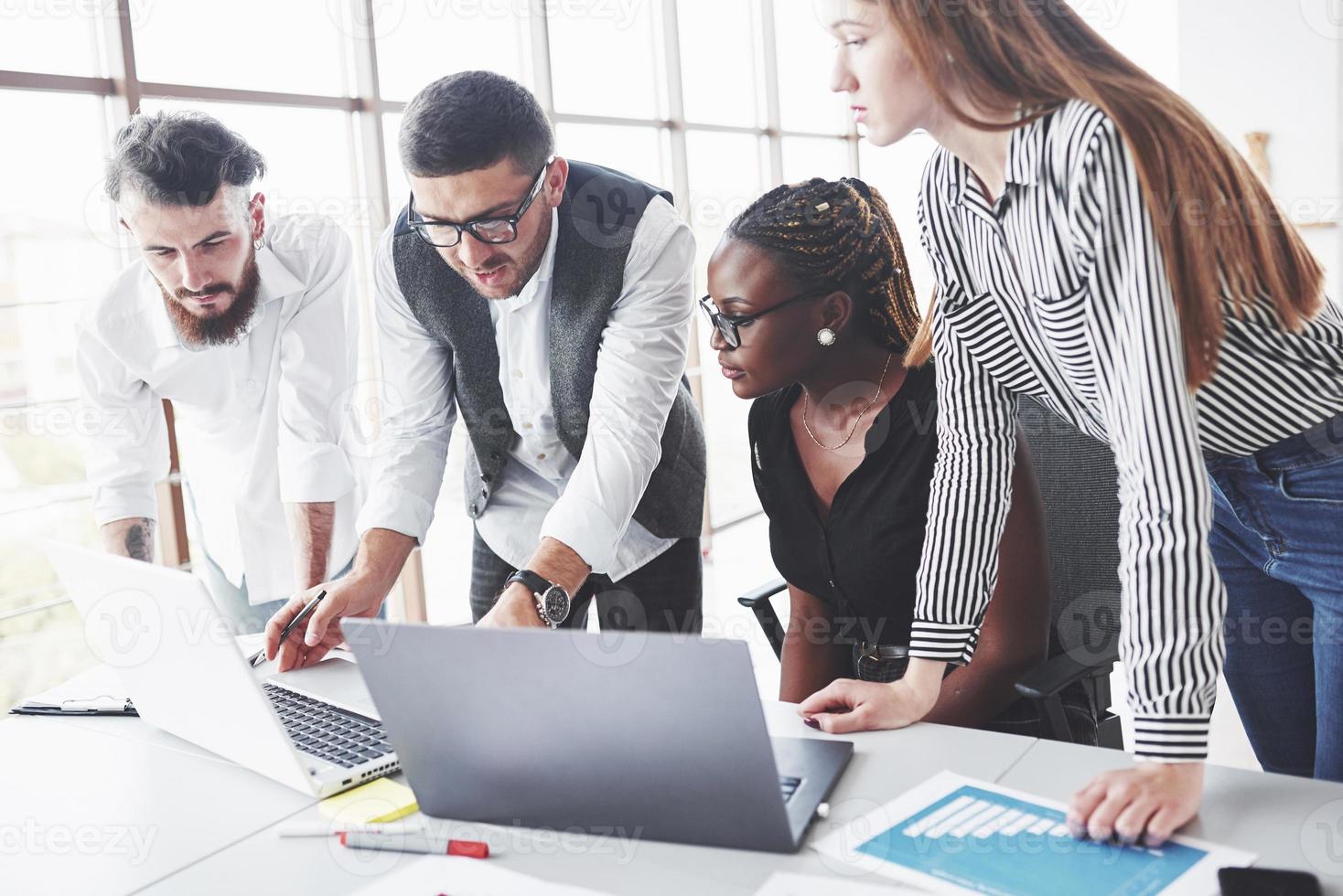 el hombre de las gafas explica la esencia de la tarea. Cuatro empleados multirraciales tienen trabajo en la oficina usando la computadora portátil en la mesa foto