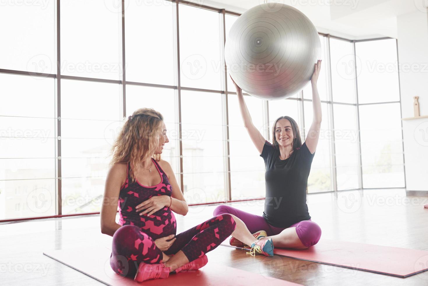 Pregnant woman sitting on the fitness mat and holding big stability ball while looking on the friend photo