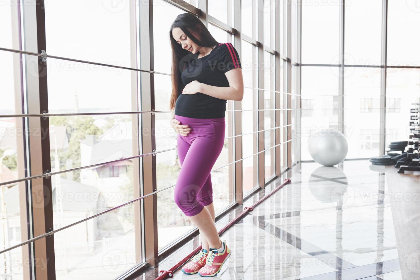 Pregnant brunette standing in the gym near the windows and keeping her hands on the belly photo