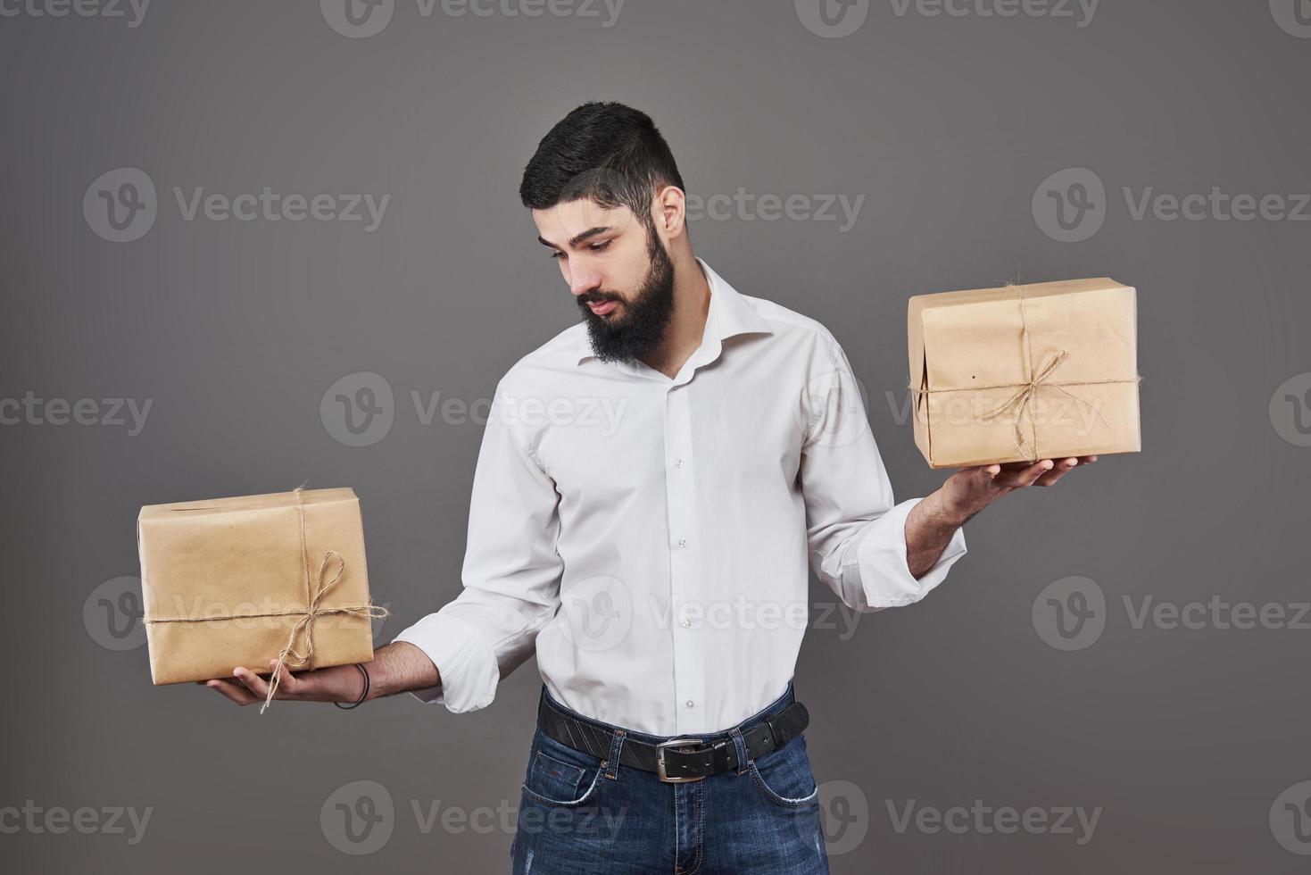 guapo chico romántico está mirando la caja y hace una elección. sosteniendo una caja de regalo de dos grandes para su pareja, sobre fondo gris. foto