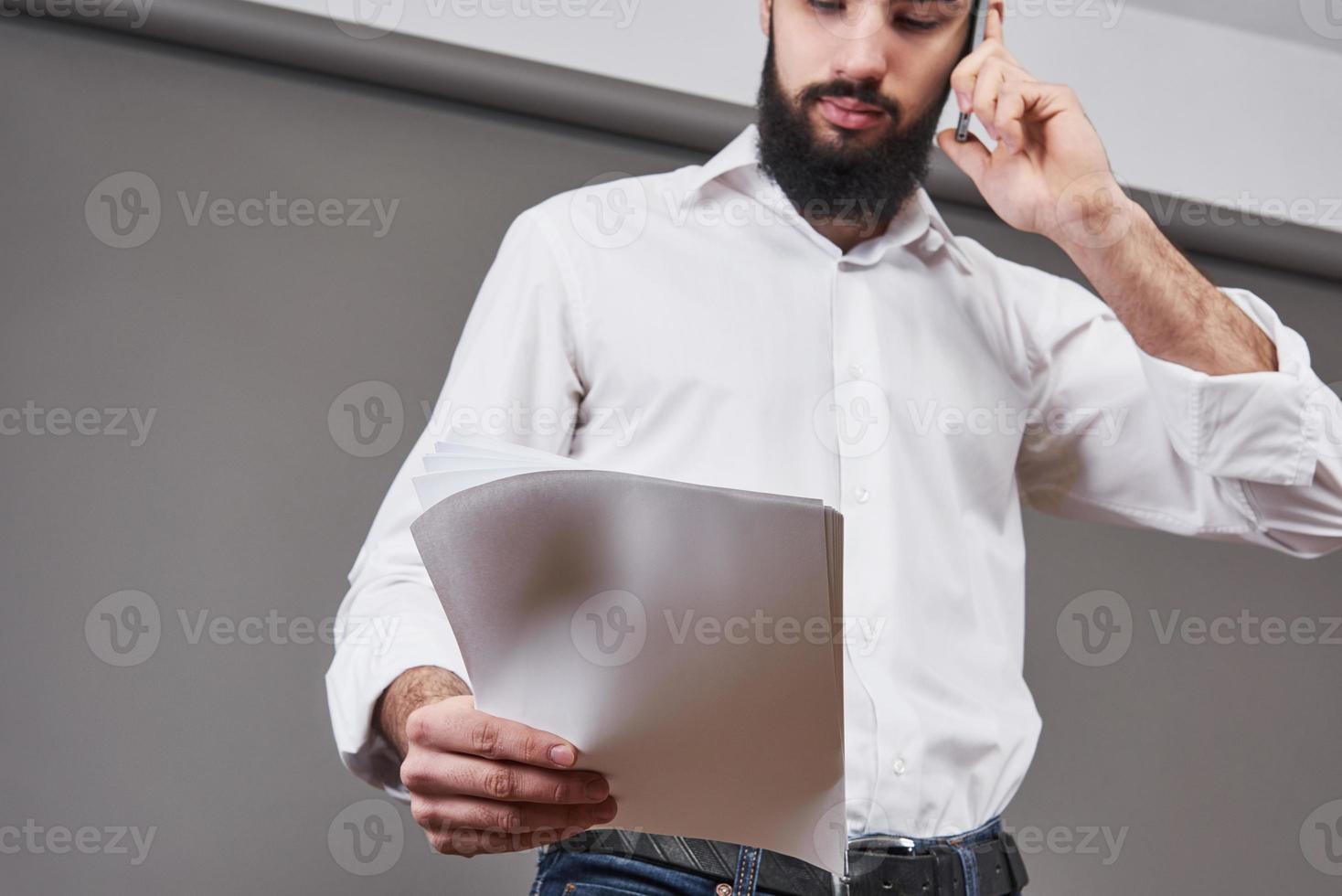 Hombre de negocios con barba en camisa blanca sosteniendo documentos y teléfono sobre fondo gris foto