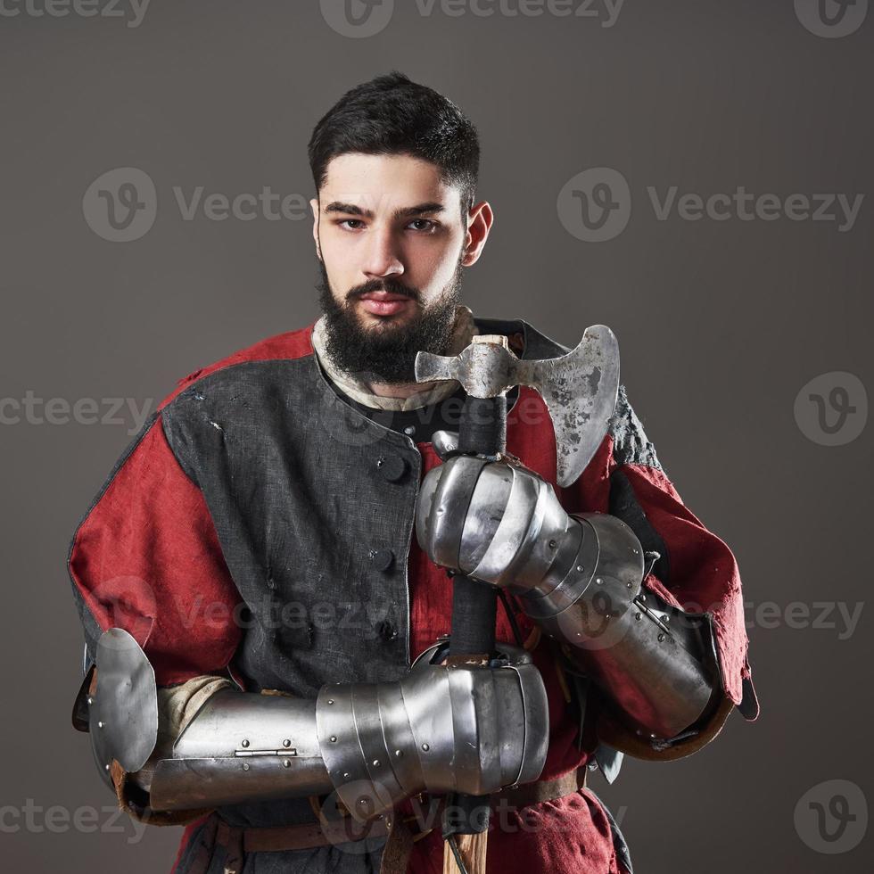 Medieval knight on grey background. Portrait of brutal dirty face warrior with chain mail armour red and black clothes and battle axe photo