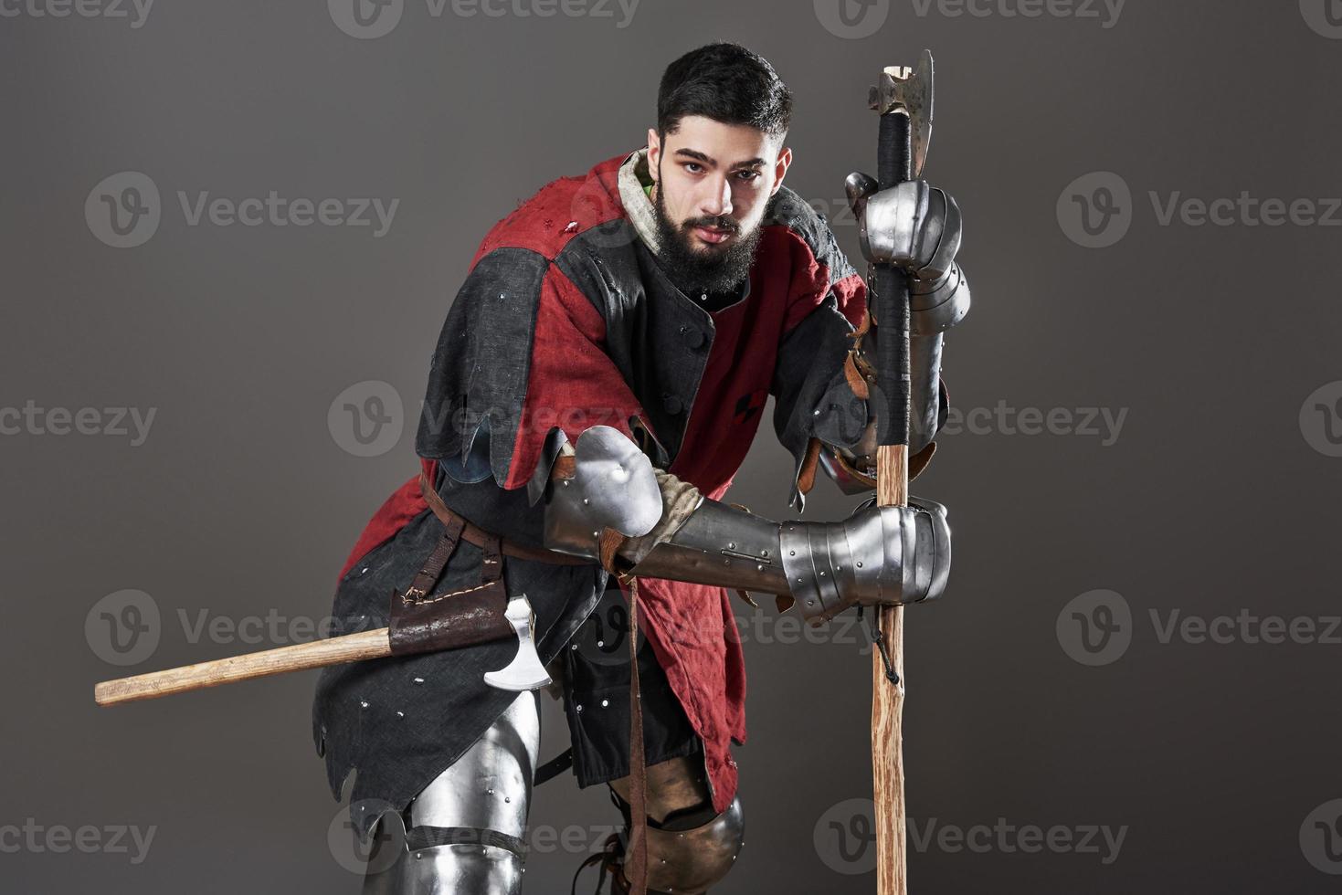 Medieval knight on grey background. Portrait of brutal dirty face warrior with chain mail armour red and black clothes and battle axe photo