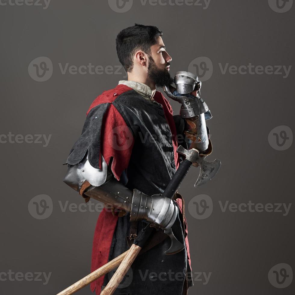 Medieval knight on grey background. Portrait of brutal dirty face warrior with chain mail armour red and black clothes and battle axe photo