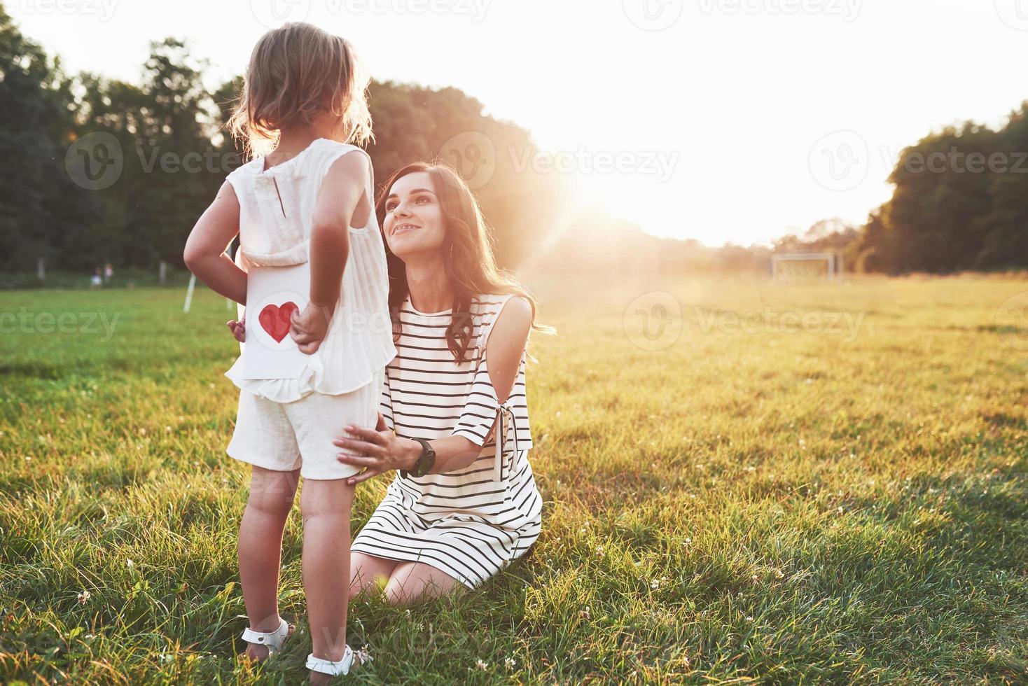 hermosa luz del sol sobre los árboles. niña esconde el libro con corazón rojo en la parte posterior de la madre sentada sonriente foto