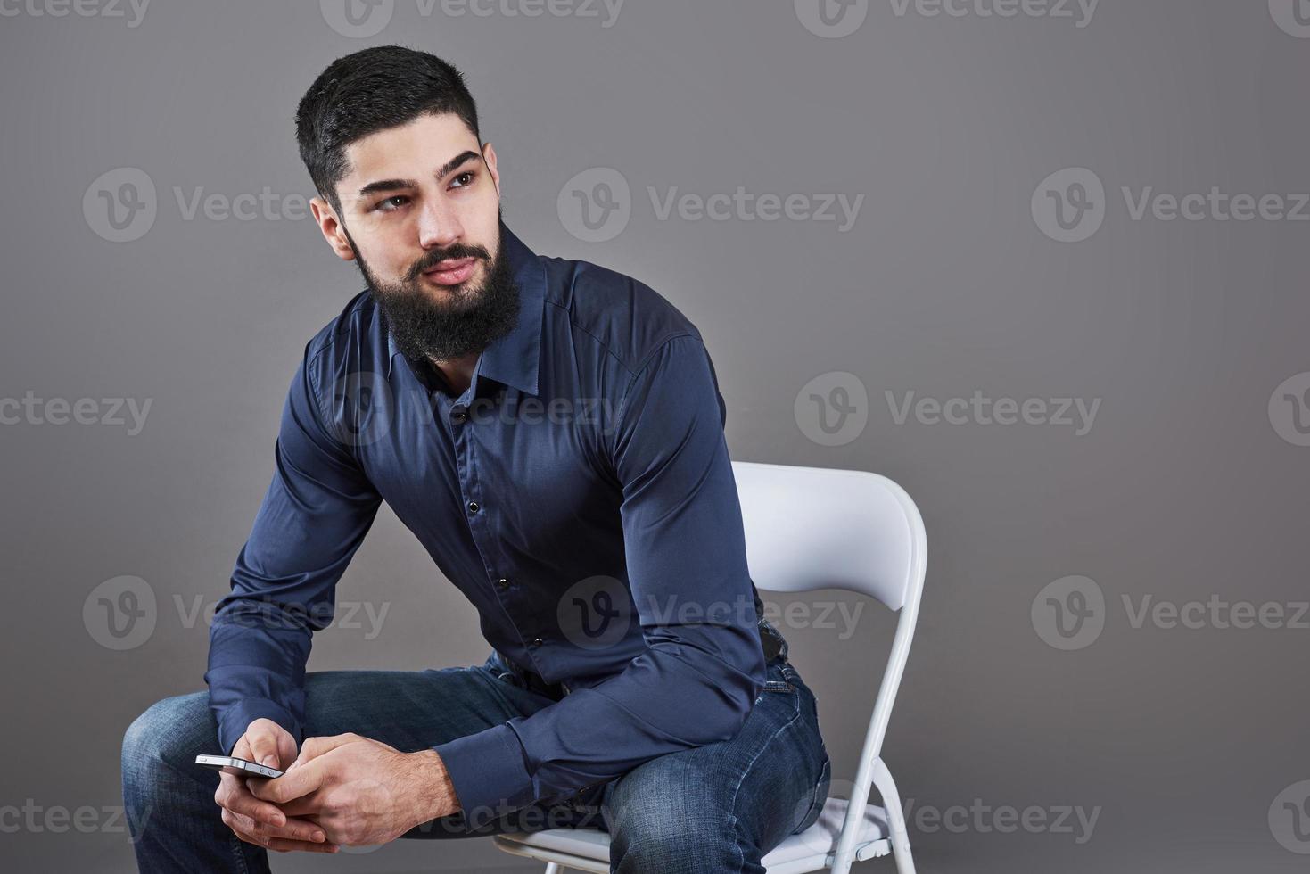 Portrait of toothy handsome bearded man sitting on a chair with phone on hands photo