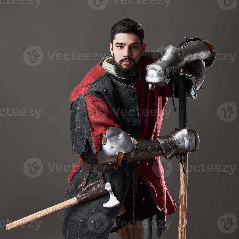 Medieval knight on grey background. Portrait of brutal dirty face warrior with chain mail armour red and black clothes and battle axe photo
