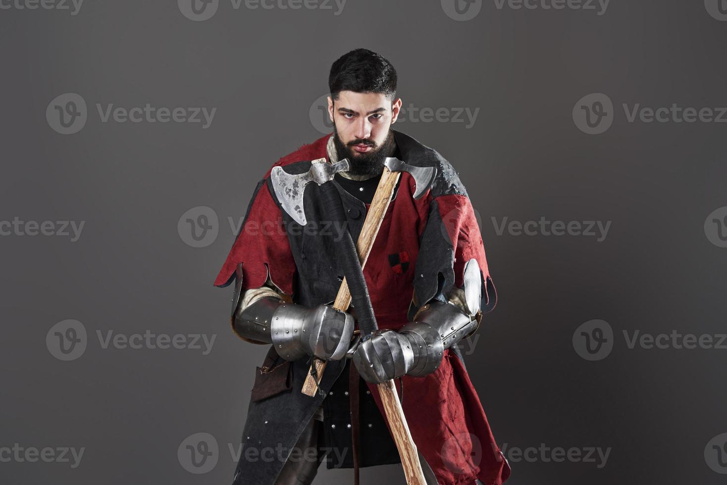 Medieval knight on grey background. Portrait of brutal dirty face warrior with chain mail armour red and black clothes and battle axe photo