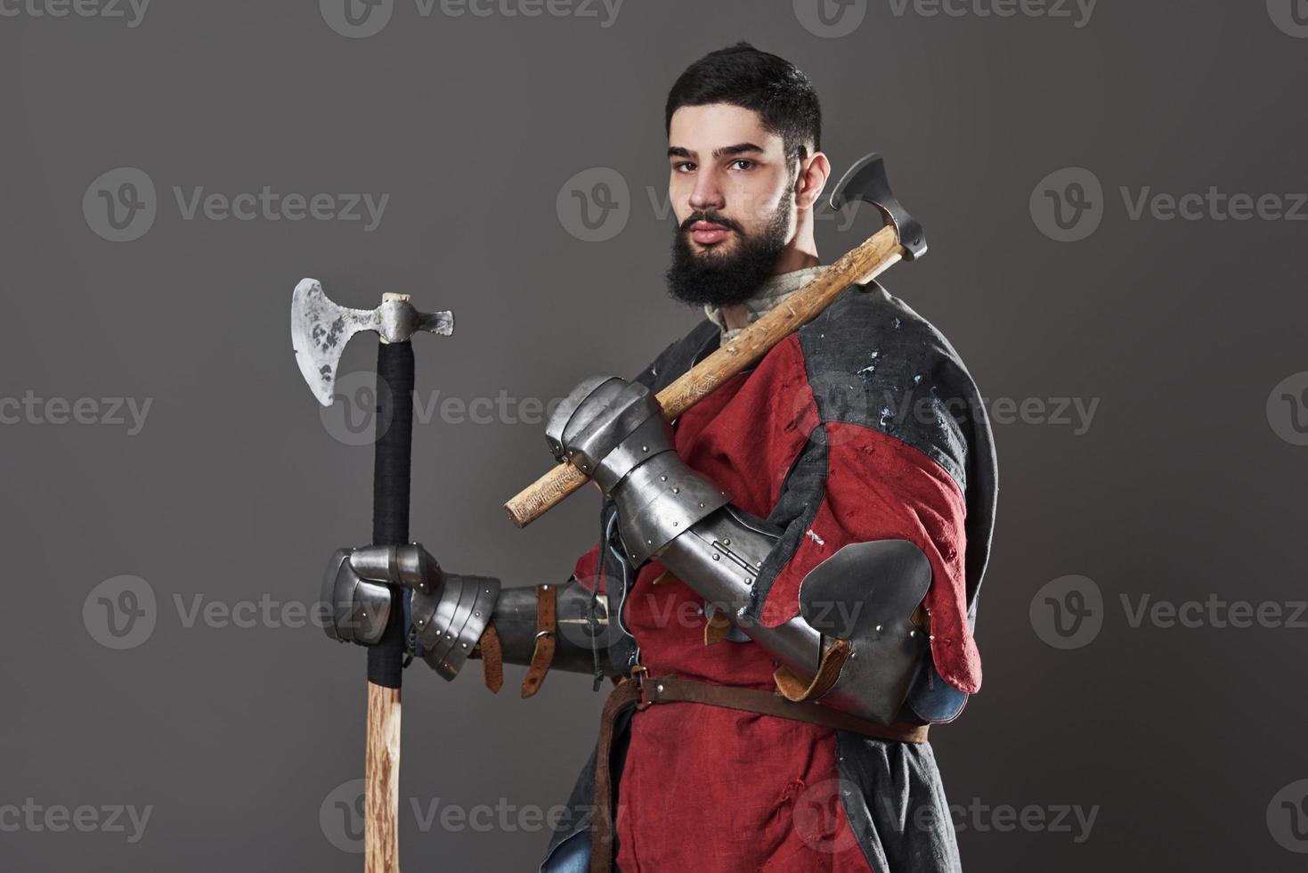 Medieval knight on grey background. Portrait of brutal dirty face warrior with chain mail armour red and black clothes and battle axe photo