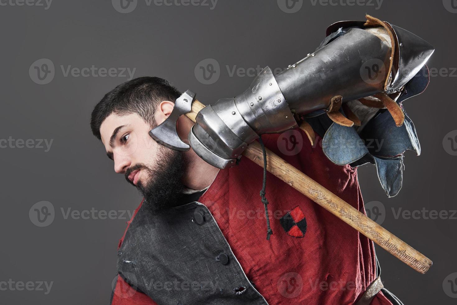 Medieval knight on grey background. Portrait of brutal dirty face warrior with chain mail armour red and black clothes and battle axe photo