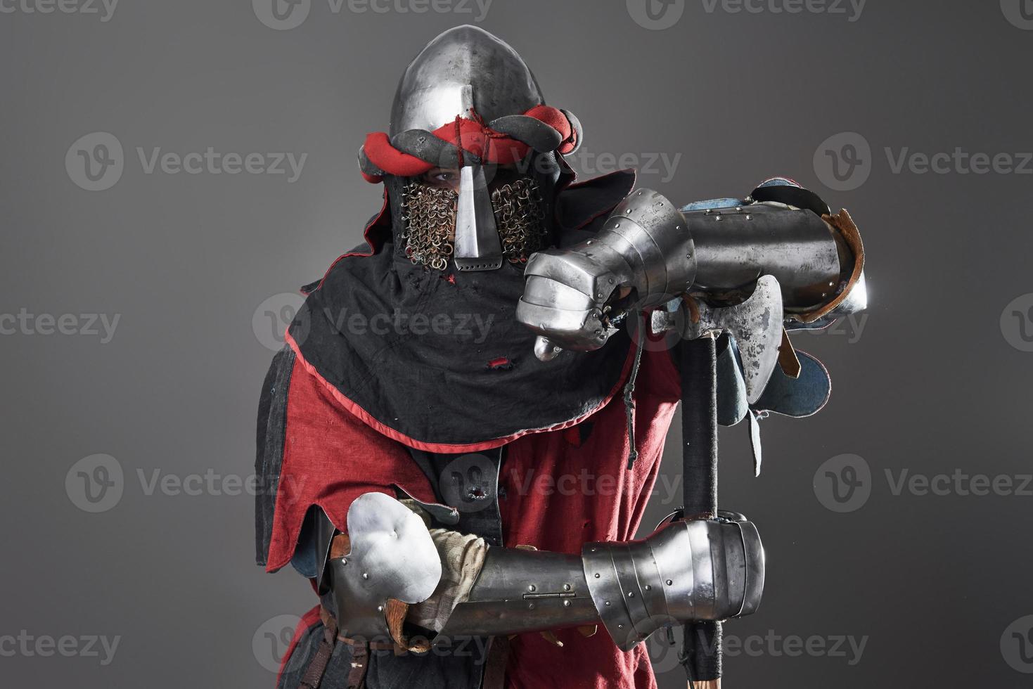 Medieval knight on grey background. Portrait of brutal dirty face warrior with chain mail armour red and black clothes and battle axe photo