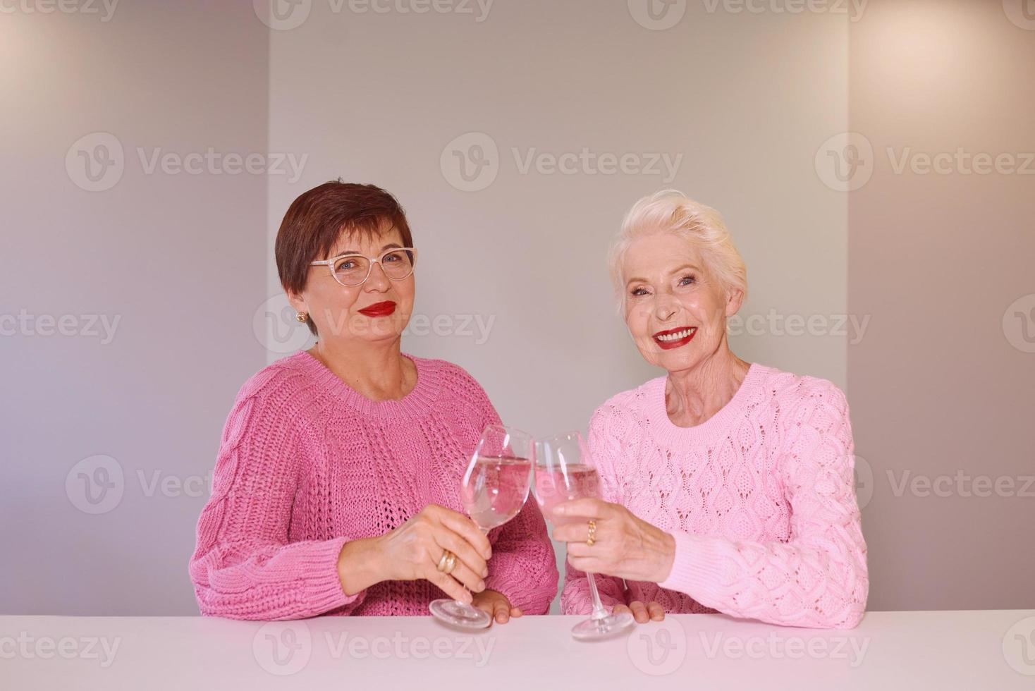 Dos mujeres mayores con estilo en suéteres de color rosa bebiendo vino rosado en la cocina moderna chismeando. amistad, hablar, chismes, eventos, relaciones, noticias, concepto de familia foto