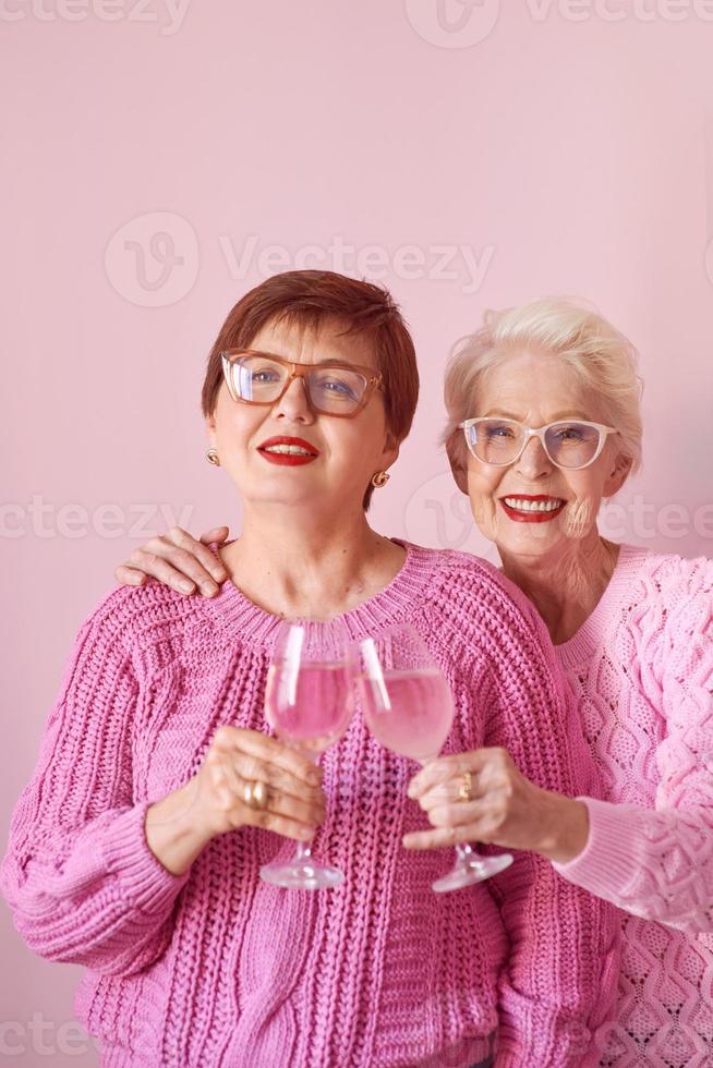 Dos mujeres mayores con estilo en suéteres de color rosa bebiendo vino rosado en la cocina moderna chismeando. amistad, hablar, chismes, eventos, relaciones, noticias, concepto de familia foto