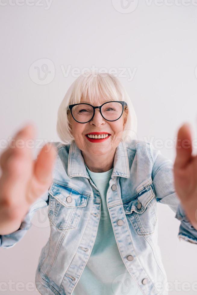 senior stylish woman with gray hair and in glasses and jeans jacket hugging you or making selfie. Love, hugs, anti age, positive vibes concept photo