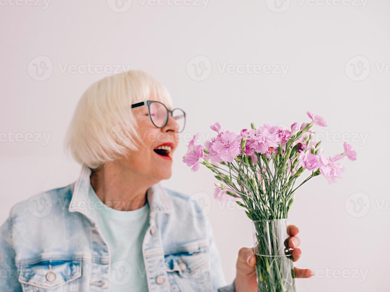 mujer con estilo senior en gafas y chaqueta de jeans huele a flores de clavel en un jarrón. flores, jubilación, profesión, concepto de hobby foto