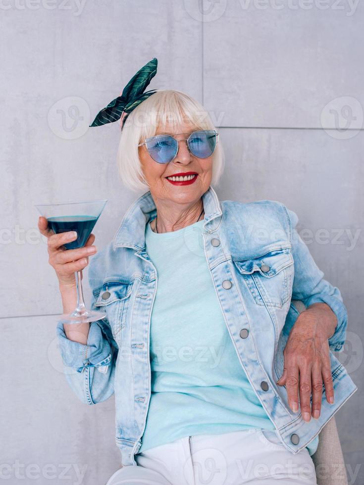 mujer elegante senior con cabello gris y gafas azules y chaqueta de mezclilla con vaso con cóctel azul. alcohol, relax, vacaciones, concepto de jubilación foto