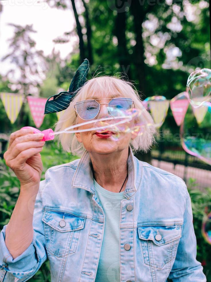 Mujer con estilo senior con cabello gris y gafas azules y chaqueta de jeans soplando burbujas al aire libre. vacaciones, fiesta, anti edad, concepto divertido foto