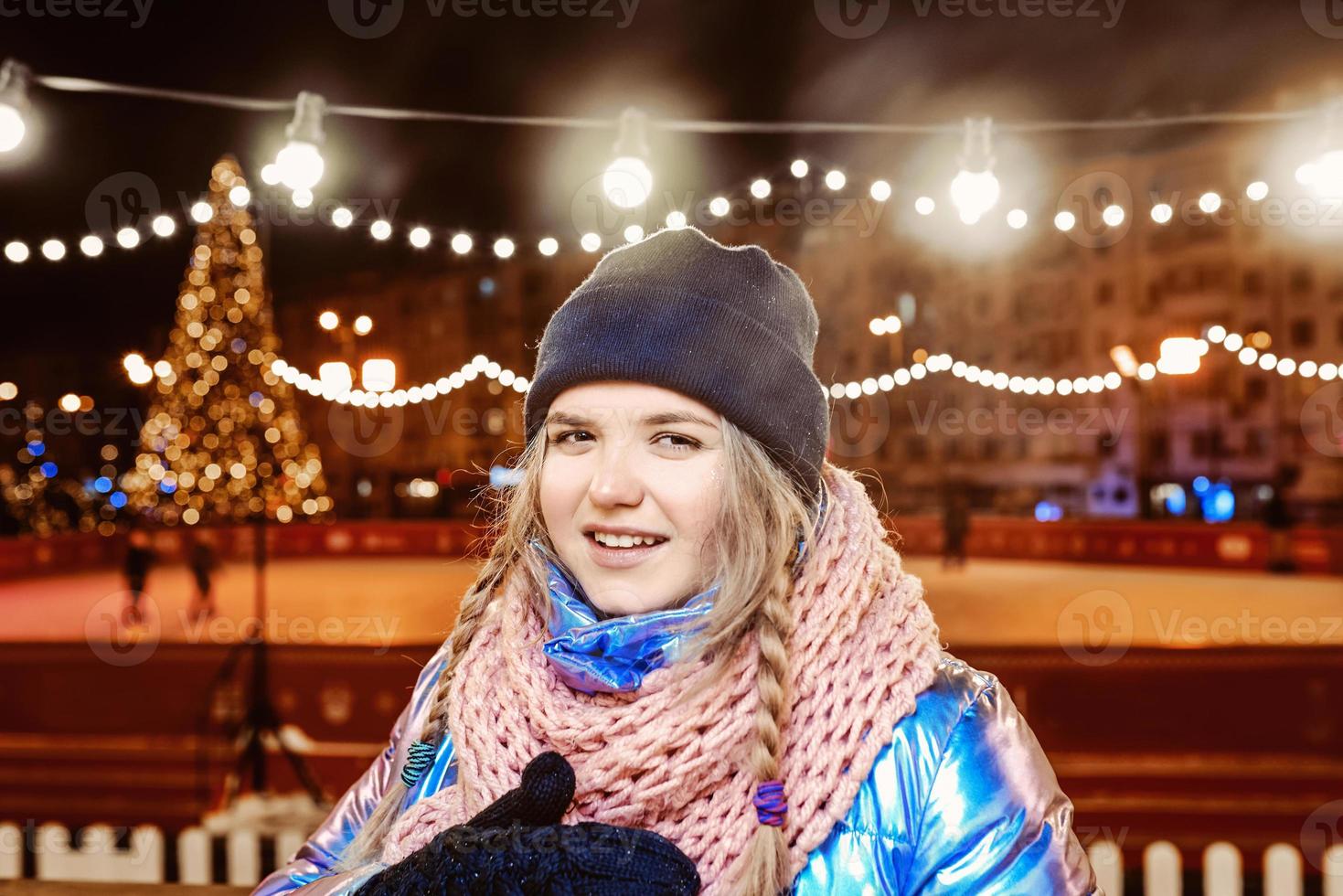 Happy smiling young Caucasian woman in scarf, hat, jacket, mittens by the outdoor rink. New year, fun, winter concept photo