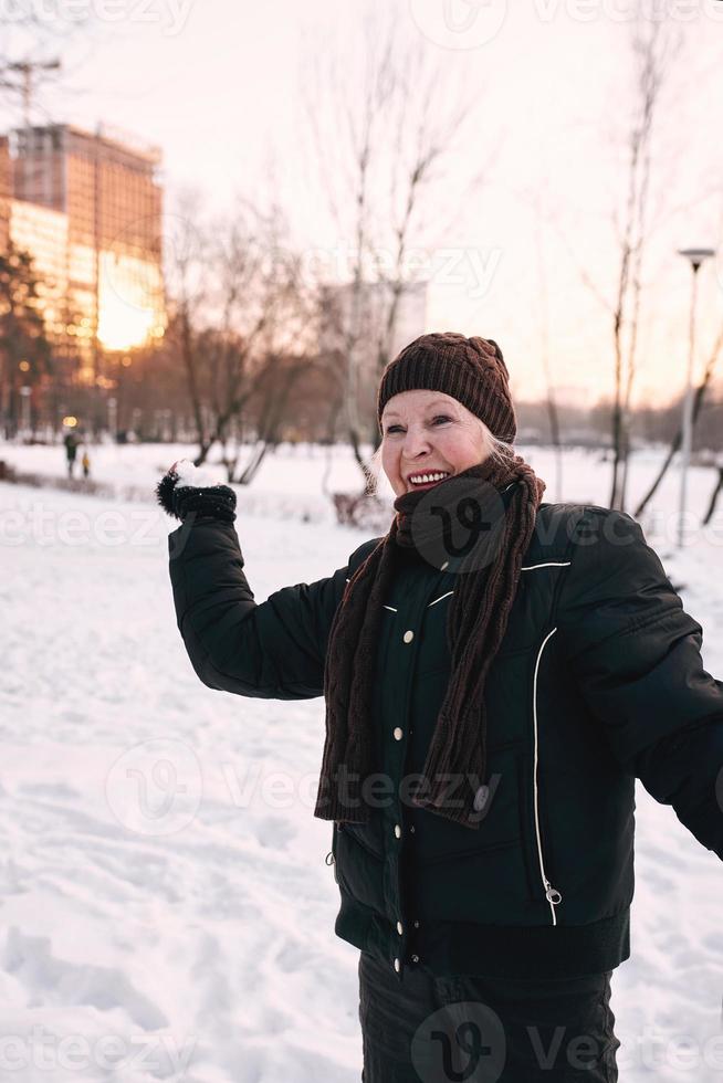 mujer mayor con sombrero y chaqueta deportiva bolas de nieve en el parque de invierno de nieve. invierno, edad, deporte, actividad, concepto de temporada foto