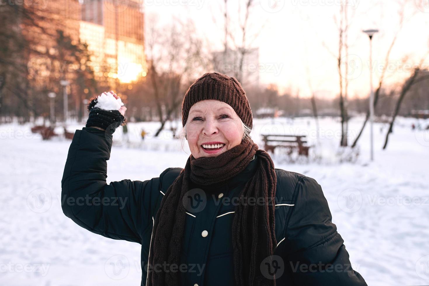 senior woman in hat and sporty jacket snowballing in snow winter park. Winter, age, sport, activity, season concept photo