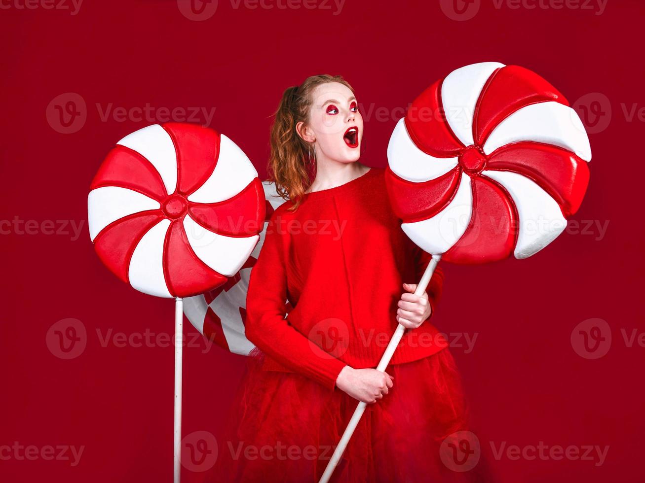 Retrato de hermosa pelirroja divertida mujer navideña sorprendida dulces sobre fondo rojo. foto