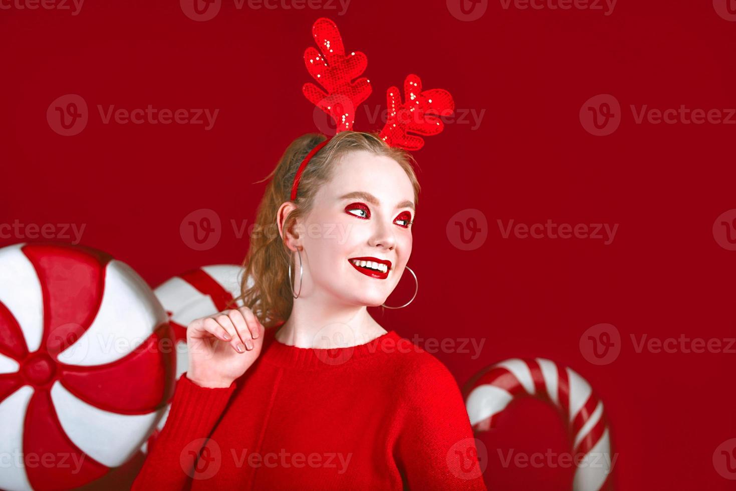 portrait of beautiful redhead funny christmas woman in deer horns with candy on the red background photo