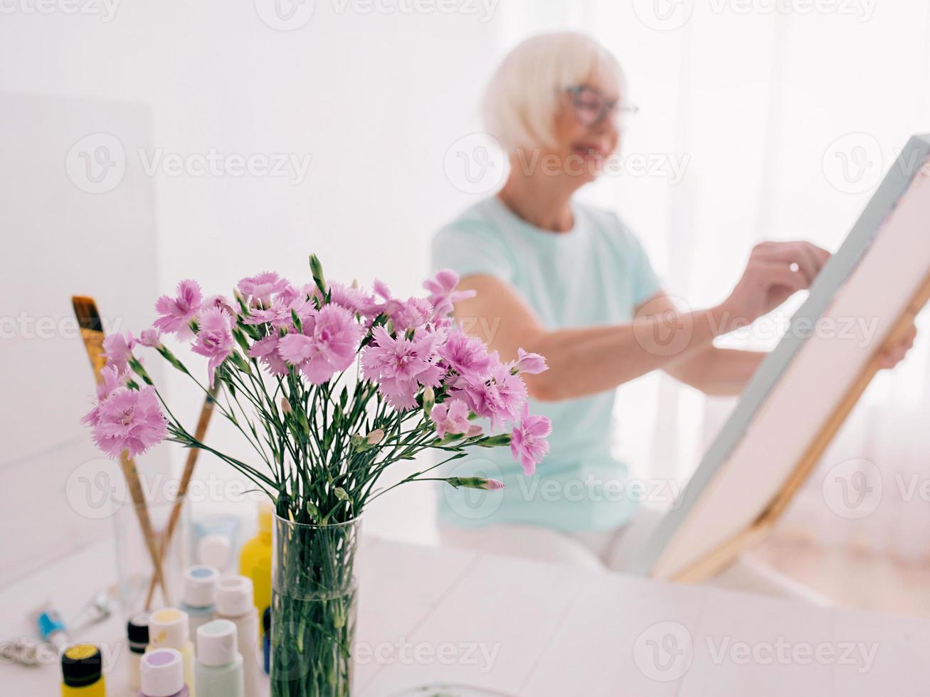 senior cheerful woman artist in glasses with gray hair painting flowers in vase. Creativity, art, hobby, occupation concept photo