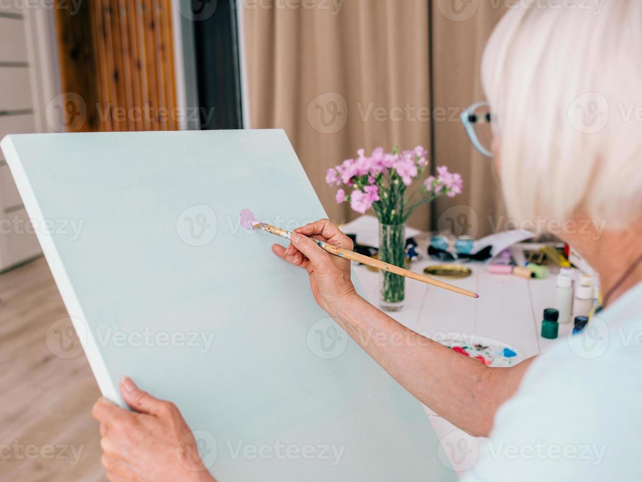 mujer alegre senior en vasos con cabello gris dibujo con lápiz flores en florero. creatividad, arte, pasatiempo, concepto de ocupación foto