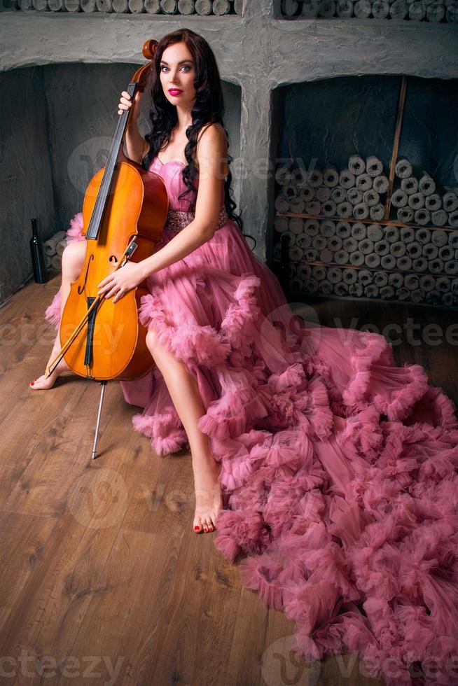 Portrait of young beautiful woman with long dark hair with cello in long pink dress in photo studio