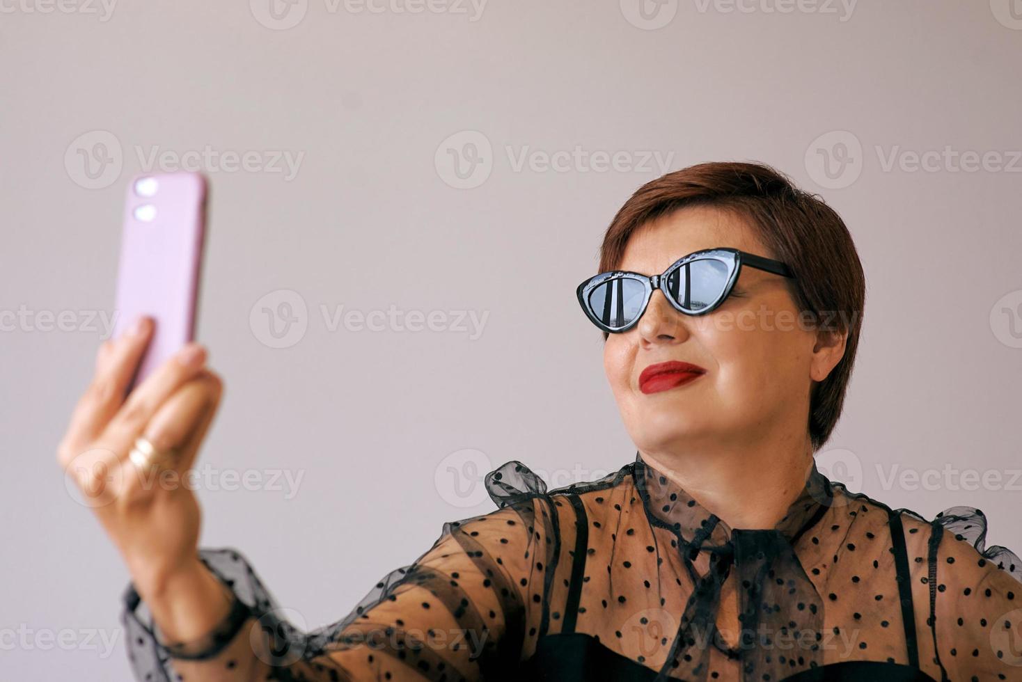 stylish mature senior woman in red blouse with cellphone video calling or making selfie. Fun, party, style, lifestyle, technology, celebration concept photo