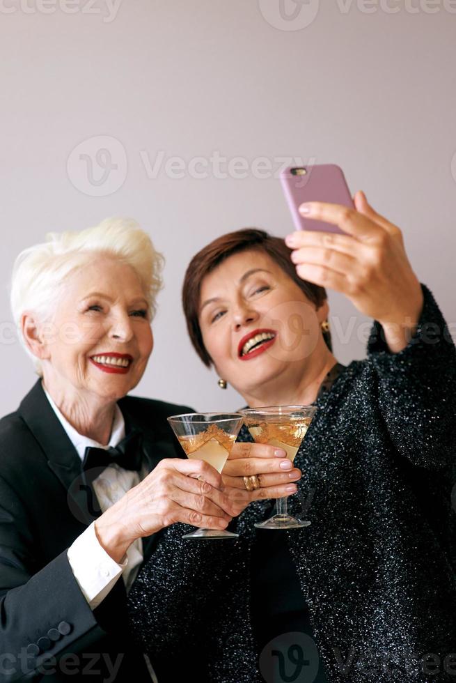 Dos hermosas mujeres mayores maduras con estilo bebiendo vino y haciendo foto selfie. diversión, fiesta, tecnología, estilo, concepto de celebración