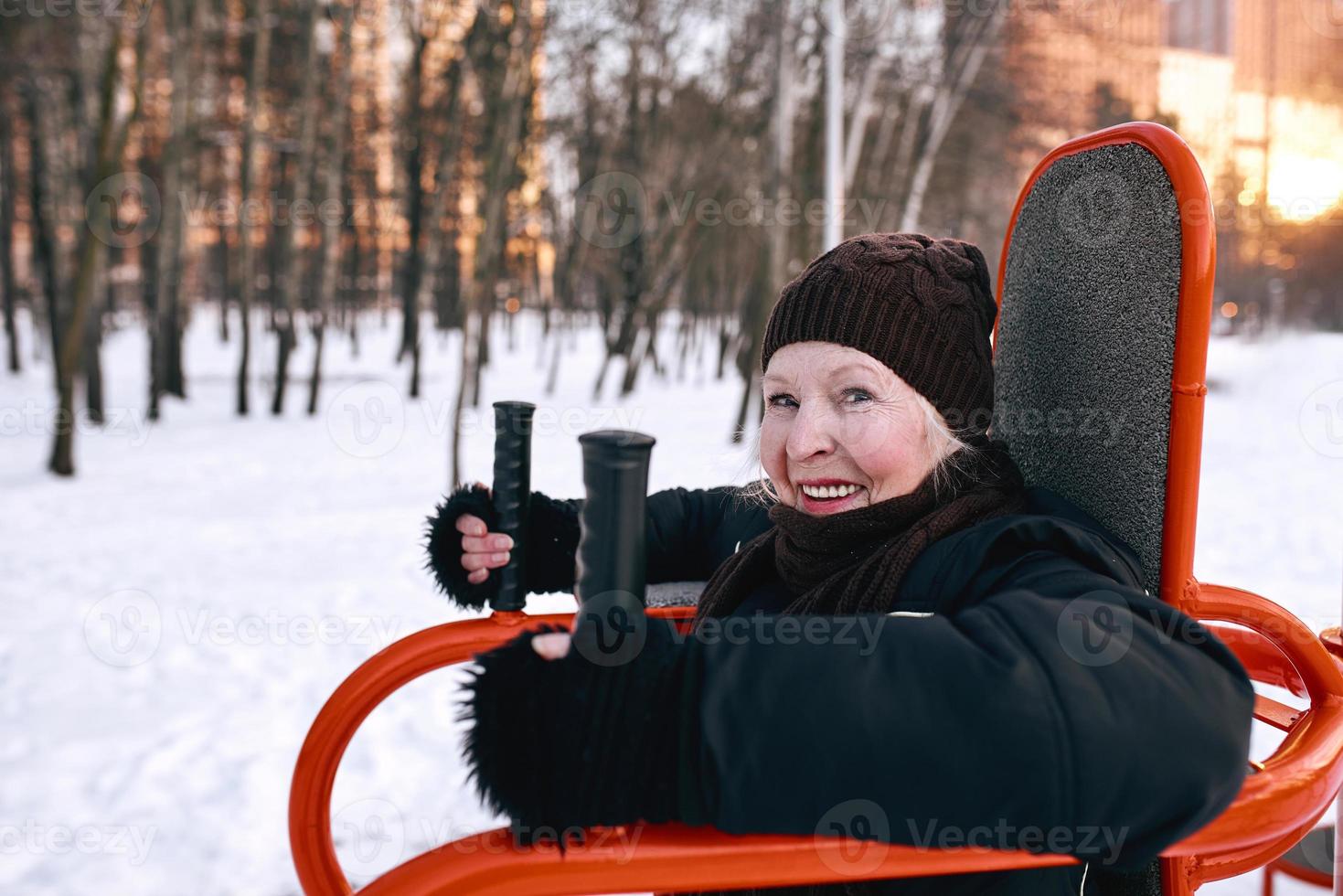 mujer mayor con sombrero y chaqueta deportiva haciendo ejercicios deportivos en el parque de invierno de nieve. invierno, edad, deporte, actividad, concepto de temporada foto