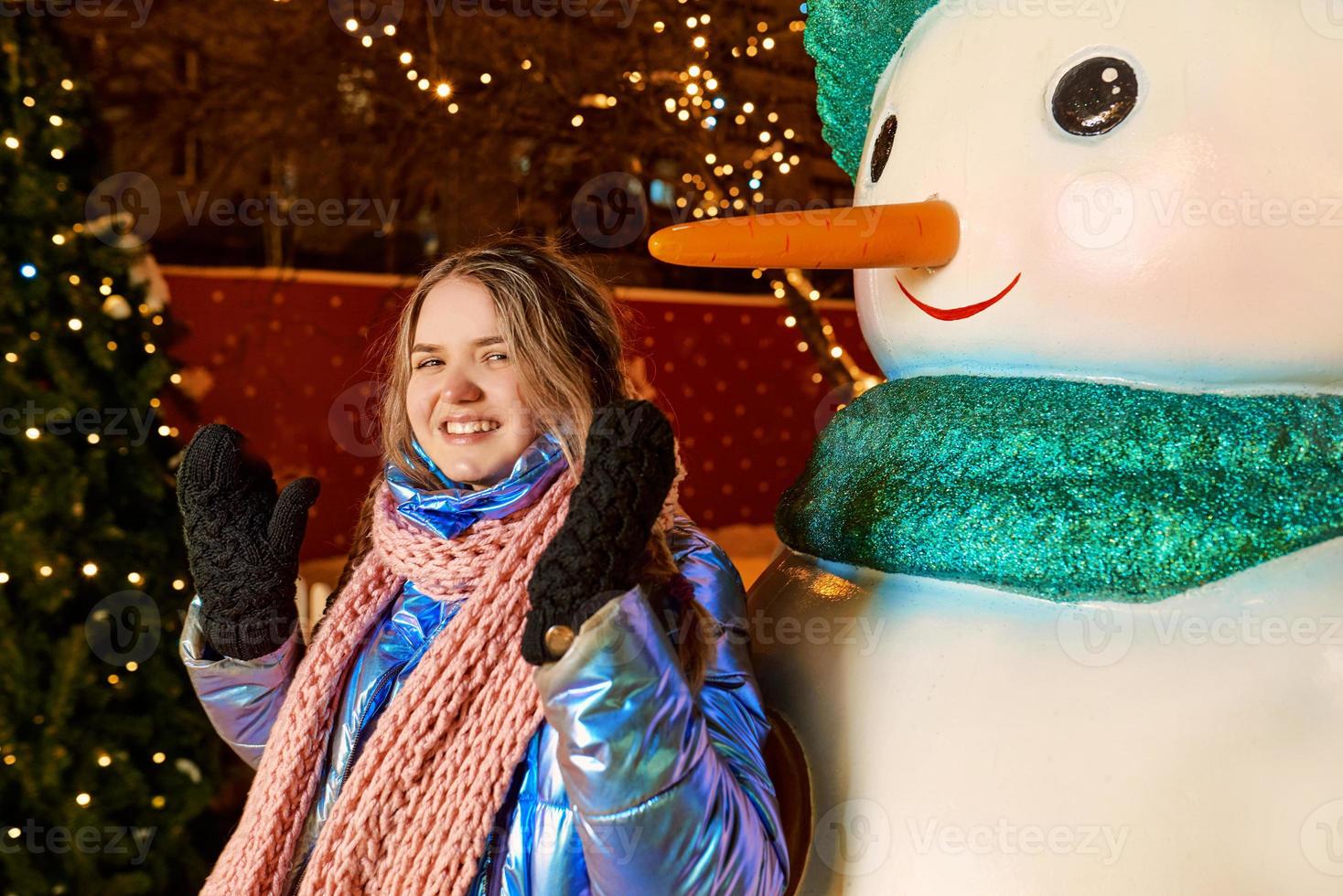 Feliz sonriente joven caucásica en bufanda, sombrero, chaqueta, guantes por el muñeco de nieve al aire libre. año nuevo, diversión, concepto de invierno. foto