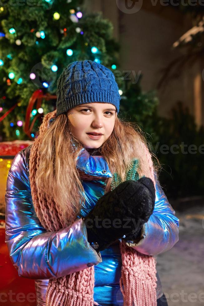 Feliz sonriente joven caucásica en bufanda, sombrero, chaqueta, guantes junto al árbol de Navidad al aire libre. año nuevo, diversión, concepto de invierno. foto